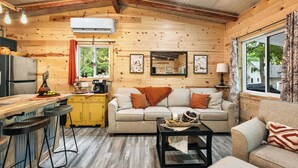 A cozy cabin interior with a wooden interior, a sofa, a coffee table, a kitchen area, and a window with curtains. - Cozy Cove Cabin The Perfect Ozark Retreat! (Camdenton)