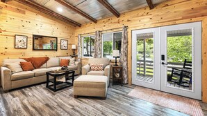 A cozy living room with a sofa, ottoman, and coffee table, wooden walls, large windows, and a door leading outside. - Cozy Cove Cabin The Perfect Ozark Retreat! (Camdenton)