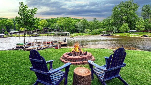 A riverside outdoor seating area with a fire pit, two Adirondack chairs, and a dock with boats. - Cozy Cove Cabin The Perfect Ozark Retreat! (Camdenton)