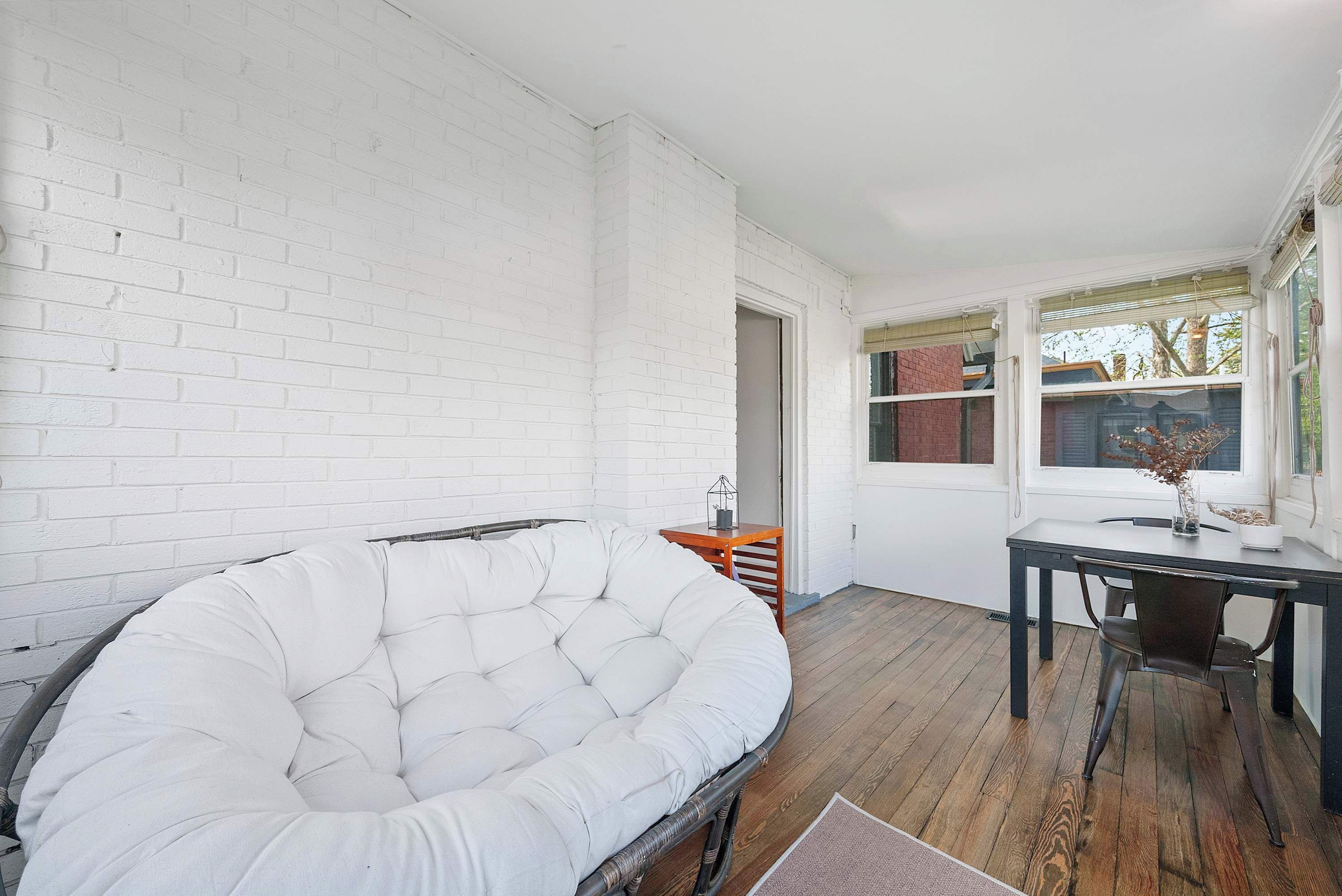 A room with a white brick wall, a white cushioned chair, a wooden table with a chair, and a window with a view of trees.