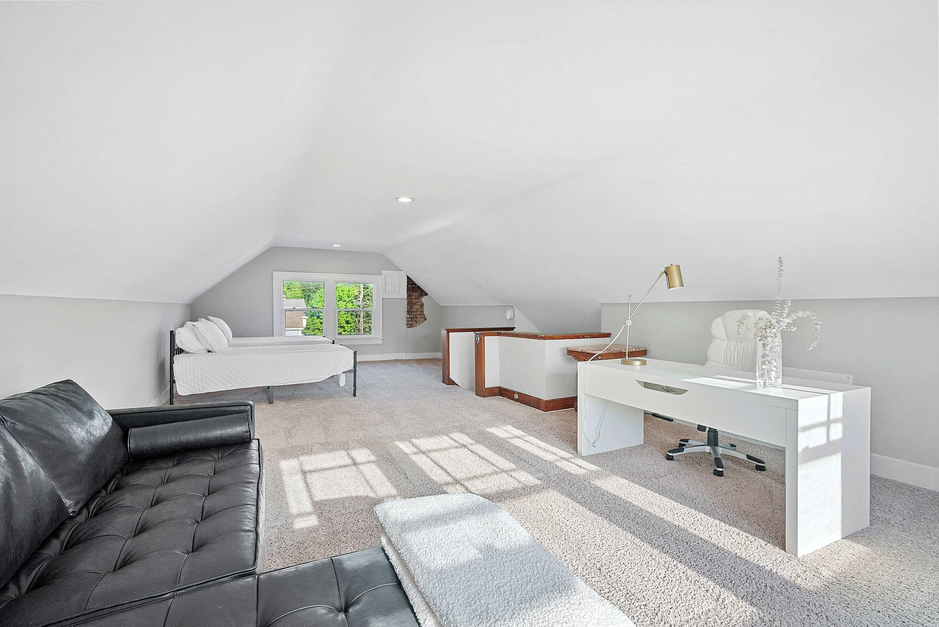 A modern loft bedroom with a sloped ceiling, a large window, a white desk, a black leather sofa, and a bed with white bedding.