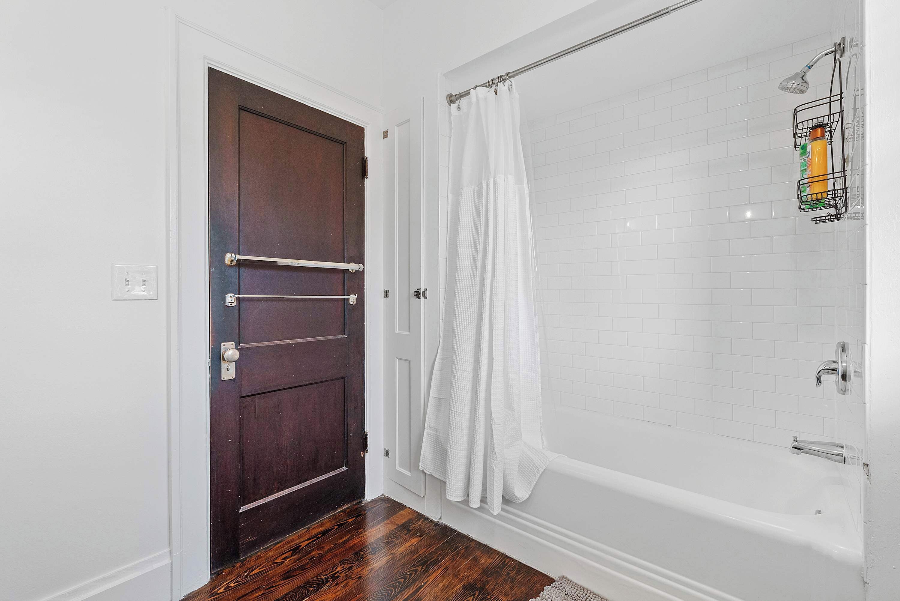 A bathroom with a wooden door, white tiled walls, a shower with a curtain, and a white bathtub.