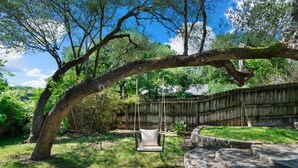 Un giardino con una sedia a dondolo sotto un albero grande, una recinzione in legno e un sentiero in pietra.