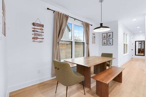 Ein Essbereich mit einem Holztisch, zwei Stühlen und einer Bank. Vor den Fenstern hängen Vorhänge und an der Wand ein Schild mit Familienweisheiten.