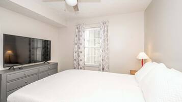 Een slaapkamer met een bed, een televisie op een dressoir, een plafondventilator en een raam met gordijnen.