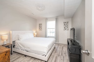 Un dormitorio con cama blanca, lámparas de noche, cómoda de madera, espejo y ventana con cortinas.