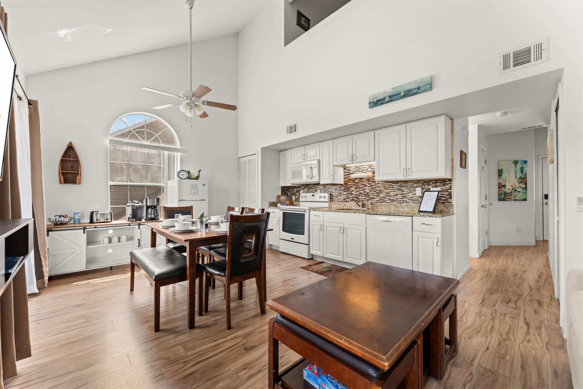 A spacious kitchen with white cabinets, a wooden dining table, and a large window.