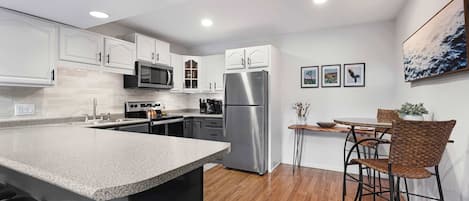 A modern kitchen with white cabinets, stainless steel appliances, a wooden dining table, and wicker chairs.
