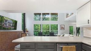 A modern kitchen with a large island, a dining area with a view of the outdoors, and a brick accent wall. - Treehouse LOZ Seclusion for 6! (Sunrise Beach)