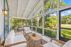 Un espacio de estar al aire libre techado con sofás blancos acolchados, una mesa de madera con jarrones decorativos y grandes ventanales que ofrecen vistas a un jardín exuberante.