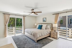 Un dormitorio con cama, mesitas de noche, ventilador de techo y ventanas grandes con cortinas.