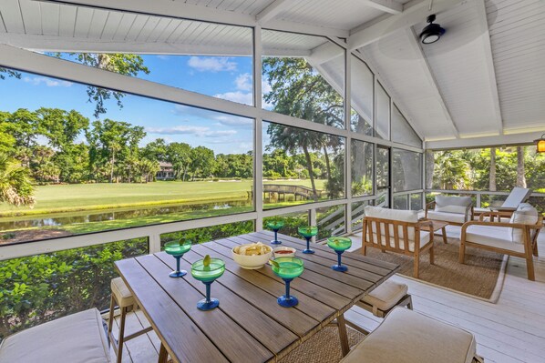 Un espacio al aire libre cubierto con una mesa de madera dispuesta para comer, rodeado de sillones de descanso y vistas a un campo de golf.
