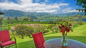 Vue depuis une pièce avec une table en verre et des chaises rouges, donnant sur un jardin luxuriant et des montagnes.