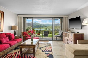 Un vaste salon doté d’une grande porte-fenêtre coulissante donnant sur un balcon, d’un téléviseur à écran plat fixé au mur et de différents types de sièges, notamment un canapé rouge, un fauteuil beige et une chaise en osier.