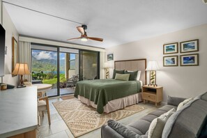A hotel room with a bed, a desk, a chair, a sofa, a ceiling fan, and a view of the outdoors.