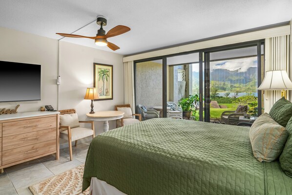 A hotel room with a bed, a desk, a chair, a television, a ceiling fan, and a view of a mountainous landscape through large windows. - (4102/03) 2Br Ground Floor Hanalei Bay Resort, Ocean View, Pool, Tennis (Princeville)