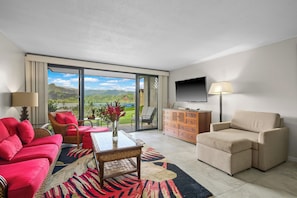 A hotel room with a red sofa, beige armchair, a TV, and a large window with a view of mountains and a lake. - (4102/03) 2Br Ground Floor Hanalei Bay Resort, Ocean View, Pool, Tennis (Princeville)