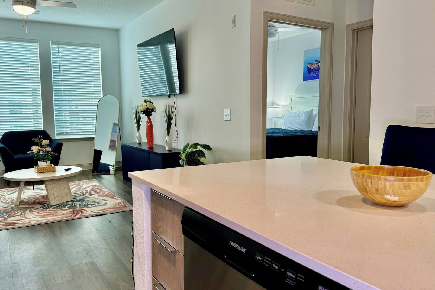 A modern kitchen with a dishwasher, a wooden bowl on the counter, and a dining area with a chair and a small table.