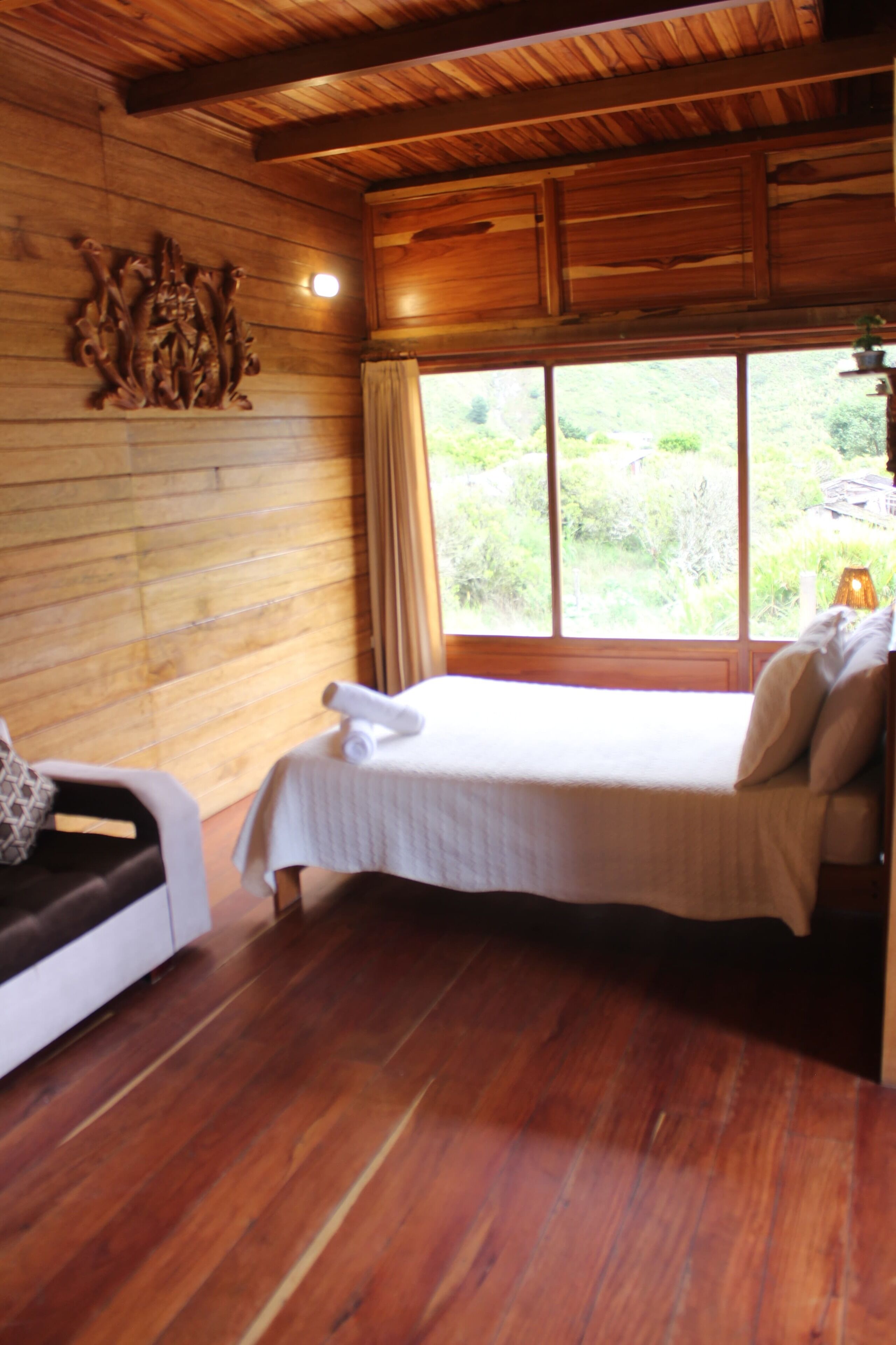 A wooden cabin room with a bed, a sofa, and a large window.