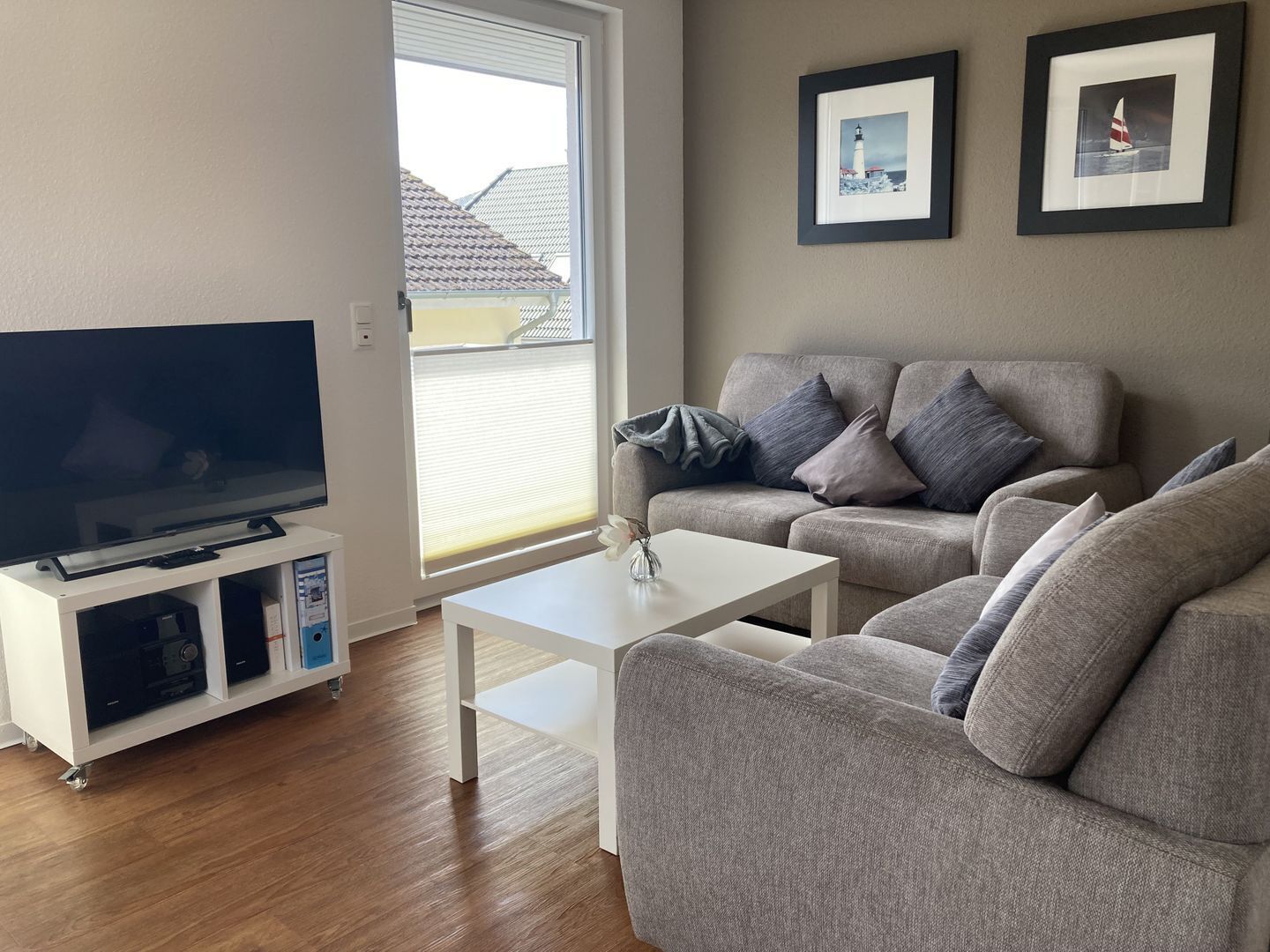 A living room with a flat-screen TV, grey sofa, white coffee table, and two framed pictures on the wall.