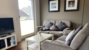 Een woonkamer met een grijze bank, een witte salontafel en een televisie op een standaard. Twee ingekaderde foto's hangen aan de muur.