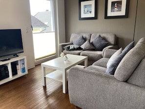 Una sala de estar con un sofá gris, una mesa de centro blanca y un televisor sobre un mueble bajo. Hay dos cuadros en la pared.