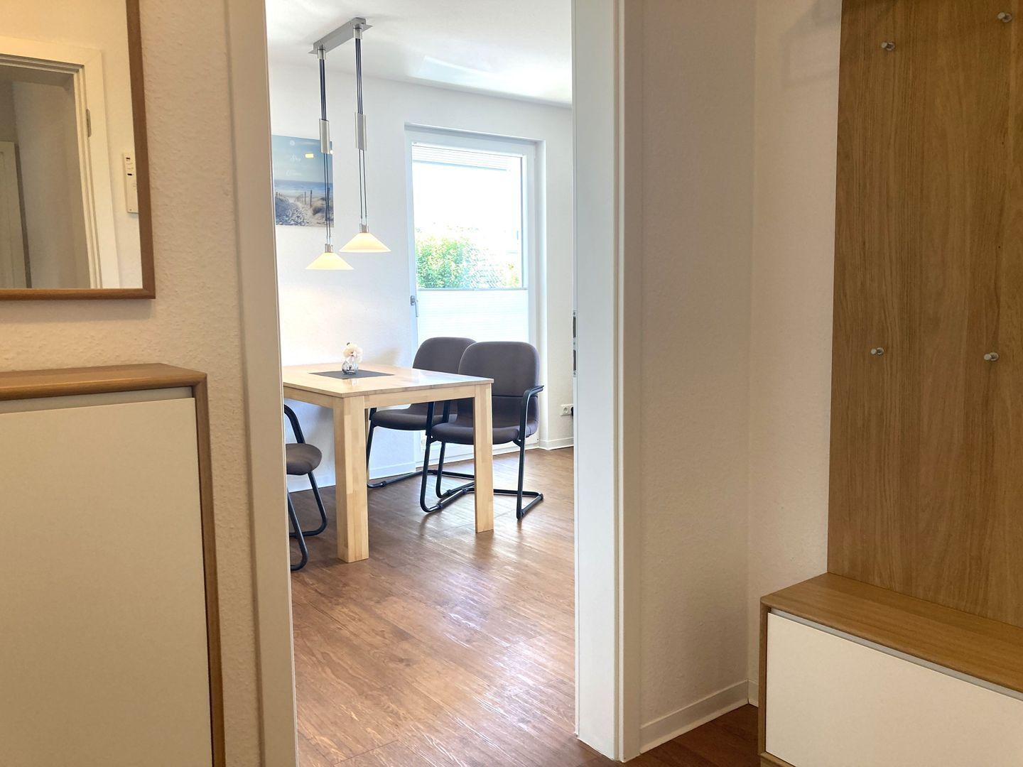 A hallway with a wooden wardrobe, a dining area with a table and chairs, and a window with a view of greenery.