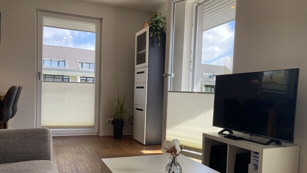 Een moderne woonkamer met een bank, een televisie op een meubel, een klein tafeltje met een vaas, en een schuifdeur naar een balkon.