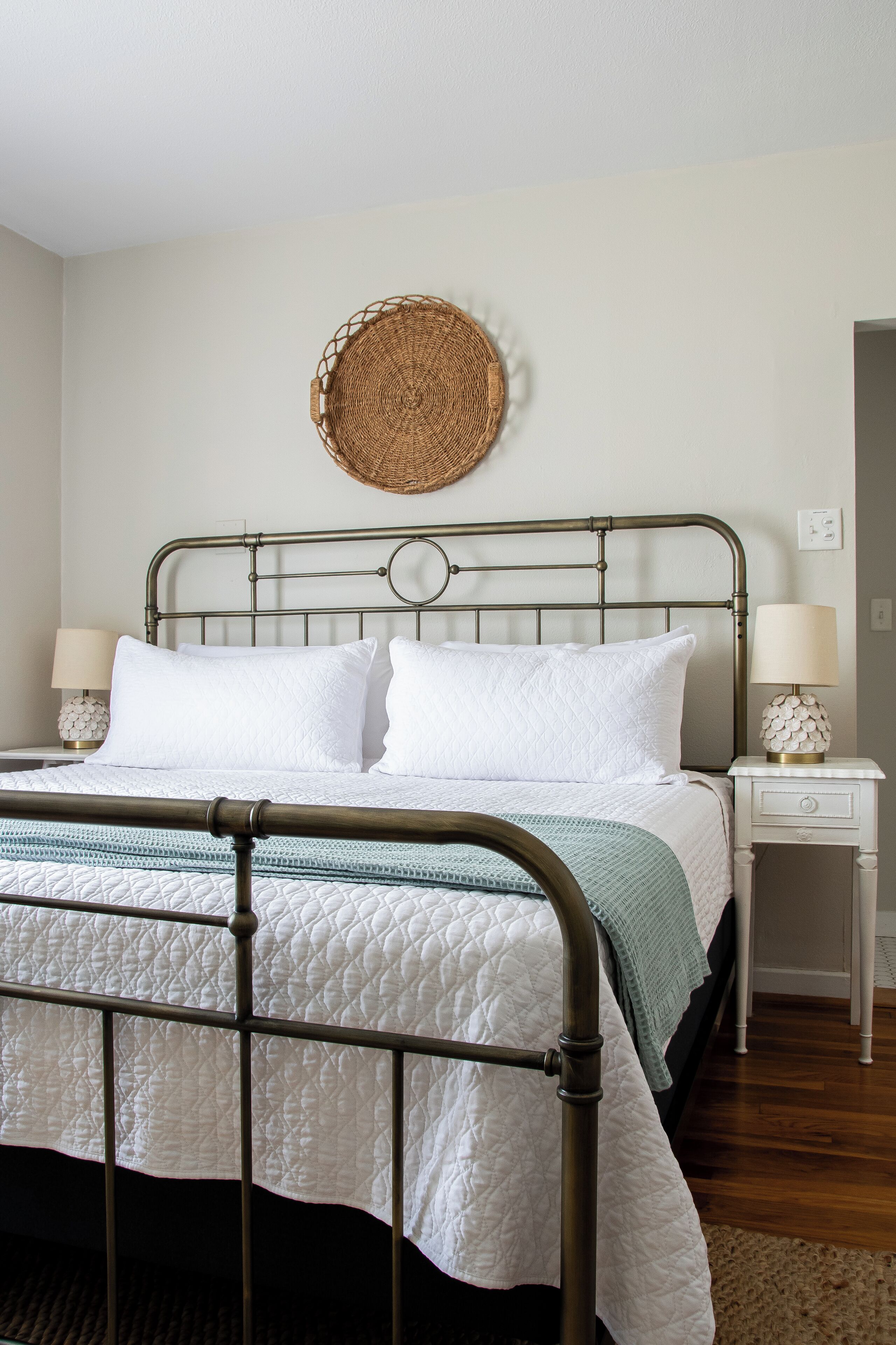 A neatly made bed with a metal frame, white bedding, and a green throw blanket. There is a bedside table with a lamp and a decorative item. A woven wall hanging is above the bed.
