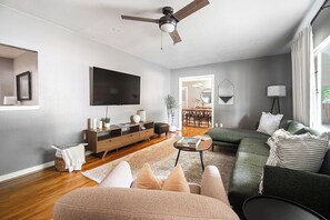 A modern living room with a flat-screen TV, a ceiling fan, a sofa, a coffee table, and a dining area visible through an open door.