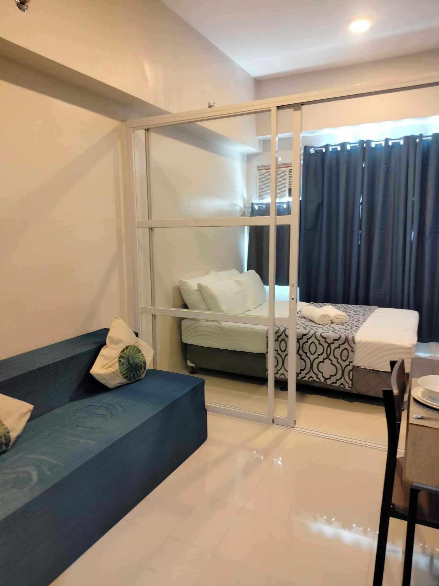 Una habitación moderna con sofá, cama con cubrecama estampado, mesa de comedor con una silla y puerta corrediza de vidrio que da a un balcón.