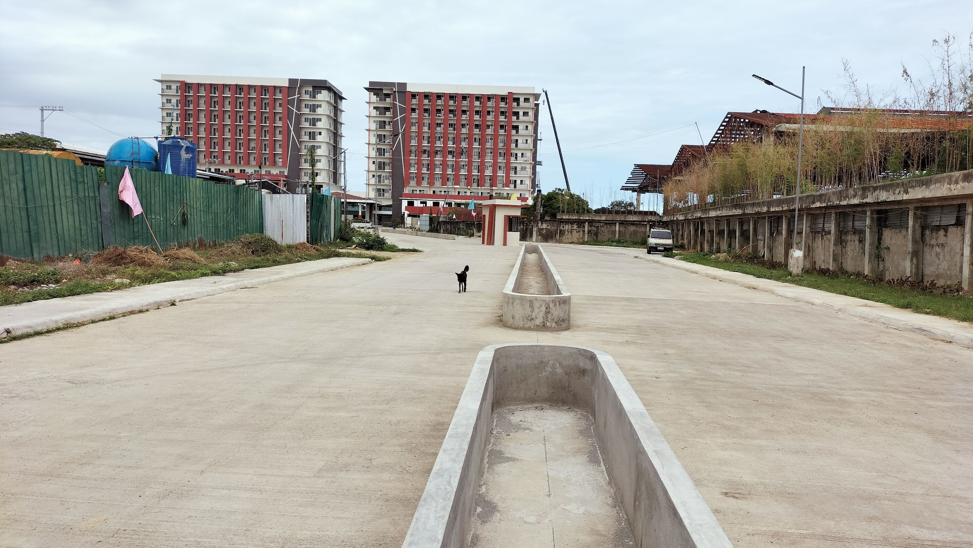 A wide, open road with a central drainage channel, flanked by green fencing and buildings with red and white facades.