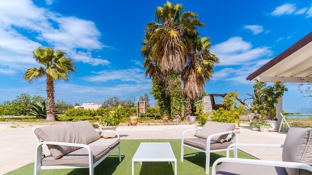 An outdoor seating area with a white table and two sofas, surrounded by palm trees and a view of the ocean.