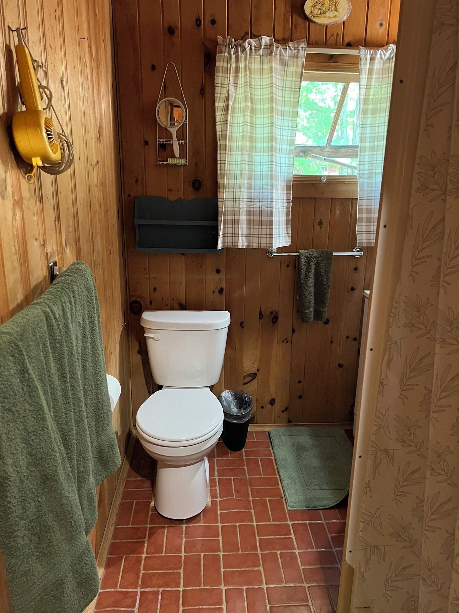 Ein Badezimmer aus Holz mit Toilette, einem grünen Handtuch, einem Mülleimer und einem Fenster mit kariertem Vorhang.