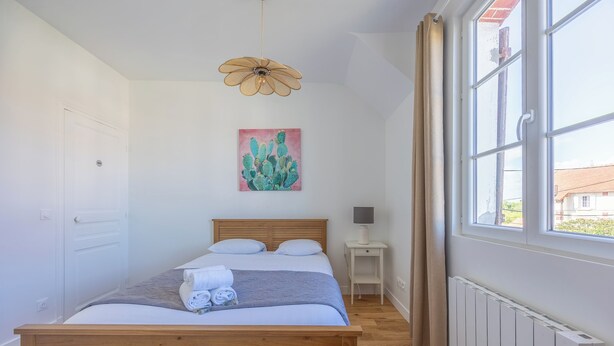 A bedroom with a wooden bed, a ceiling fan, a bedside table with a lamp, a window with curtains, and a cactus painting on the wall. - La Maison du Fleuve - Welkeys (Dives-sur-Mer)