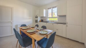 A modern kitchen with white cabinets, a dining table set for four, and a window with a view of a house. - La Maison du Fleuve - Welkeys (Dives-sur-Mer)