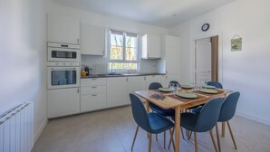 A modern kitchen with white cabinets, a wooden dining table with blue chairs, and a window providing natural light. - La Maison du Fleuve - Welkeys (Dives-sur-Mer)
