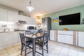 Une cuisine moderne avec des meubles blancs, une table à manger avec des chaises, un réfrigérateur, un téléviseur fixé au mur et un luminaire au-dessus de la table.