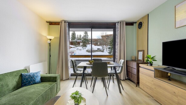 A modern living room with a sofa, dining table, chairs, a TV, and a view of a snowy landscape through large windows.