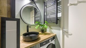 A compact bathroom with a wooden vanity, a black basin, a potted plant, a round mirror, and a front-loading washing machine.