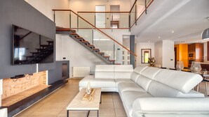 A modern living room with a large flat-screen TV, a white leather sofa, a wooden coffee table, and a staircase with glass railings. - Villa "Costacabana - Marcella" with Private Pool, Private Terrace & Wi-Fi (Lloret de Mar)
