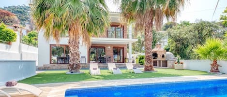 A modern house with a swimming pool, palm trees, and a barbecue area.