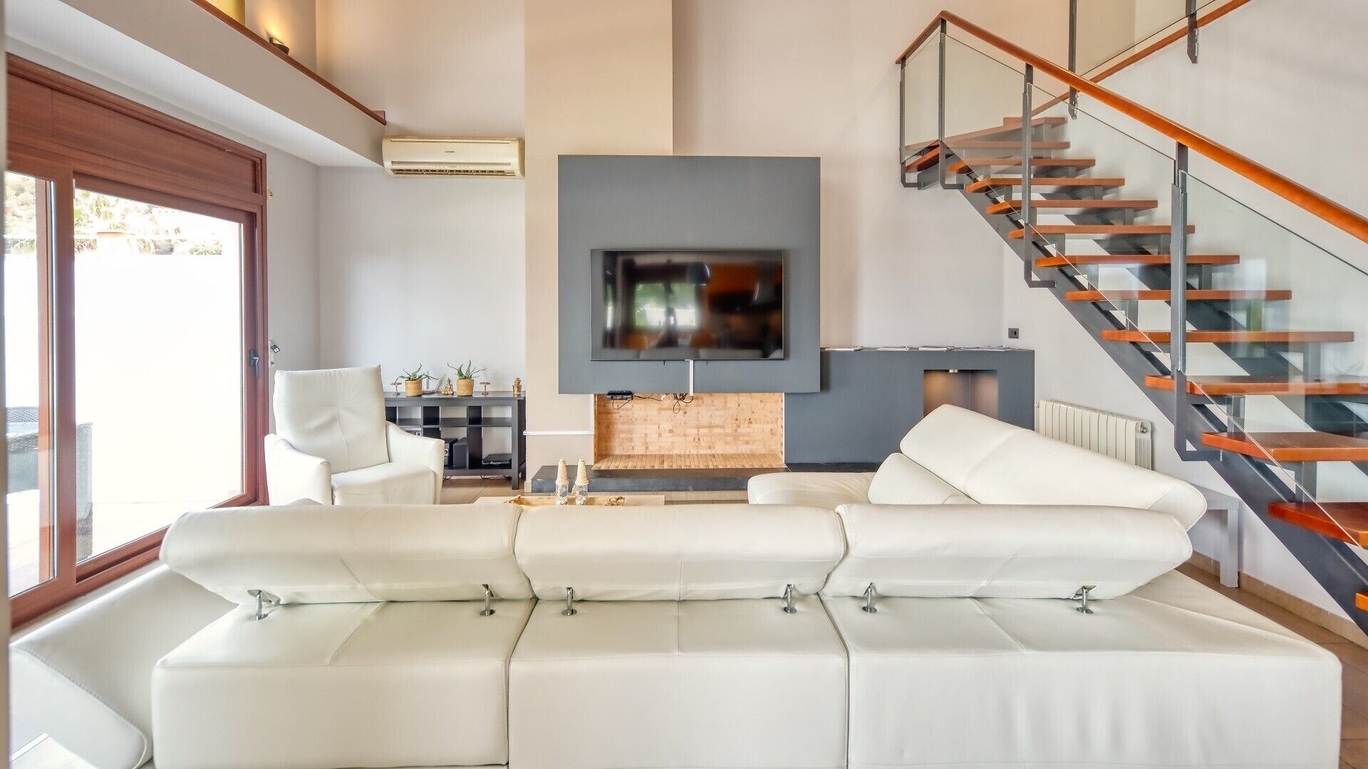 A modern living room with a sectional sofa, a flat-screen TV, a fireplace, and a staircase.