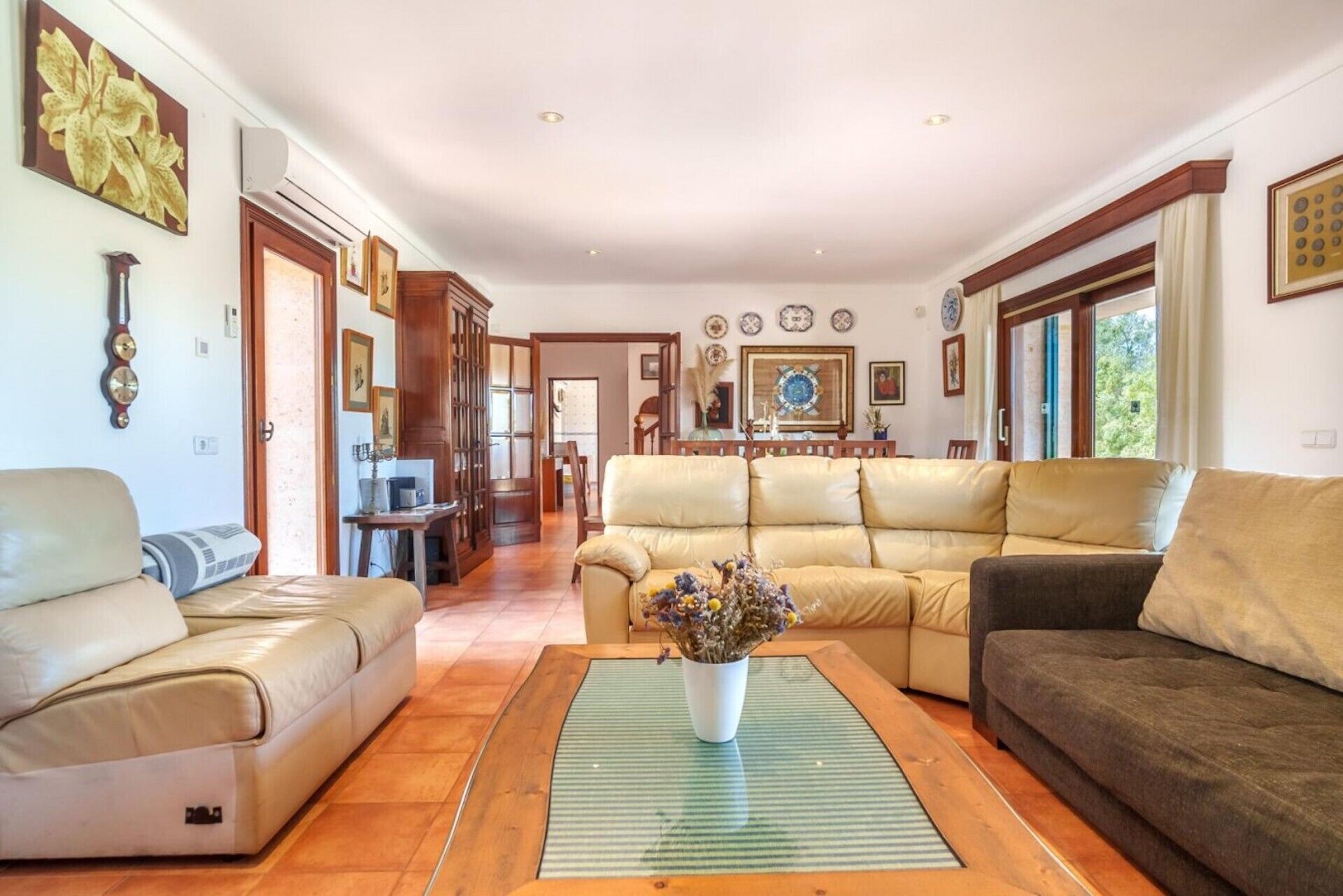 A spacious living room with a beige sofa, a dark brown sofa, and a white armchair. A wooden coffee table with a vase of flowers is in the center.