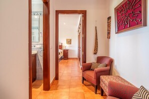 Un couloir mène à une chambre qui contient un lit, une table de chevet et une lampe. Le salon est aménagé avec un canapé rouge et un pouf à motifs.