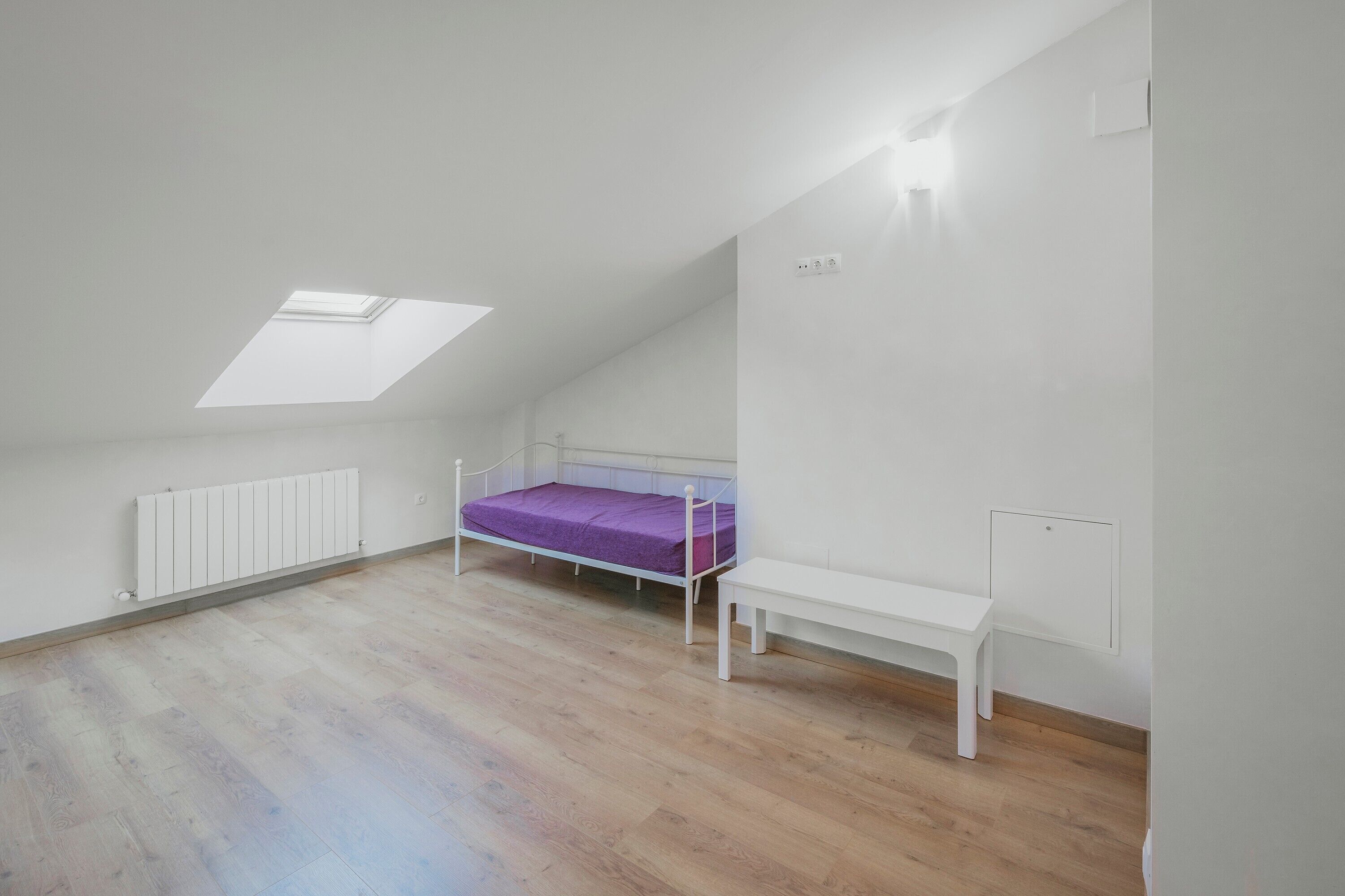A room with a sloped ceiling, a skylight, a bed with a purple mattress, a white bench, and a radiator.