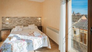 A bedroom with a large bed, a nightstand, a radiator, and a view of buildings through a window. - Tradition Rural El Tío Ricardo Country House – Private Pool & Garden in Segovia (NIEVA)