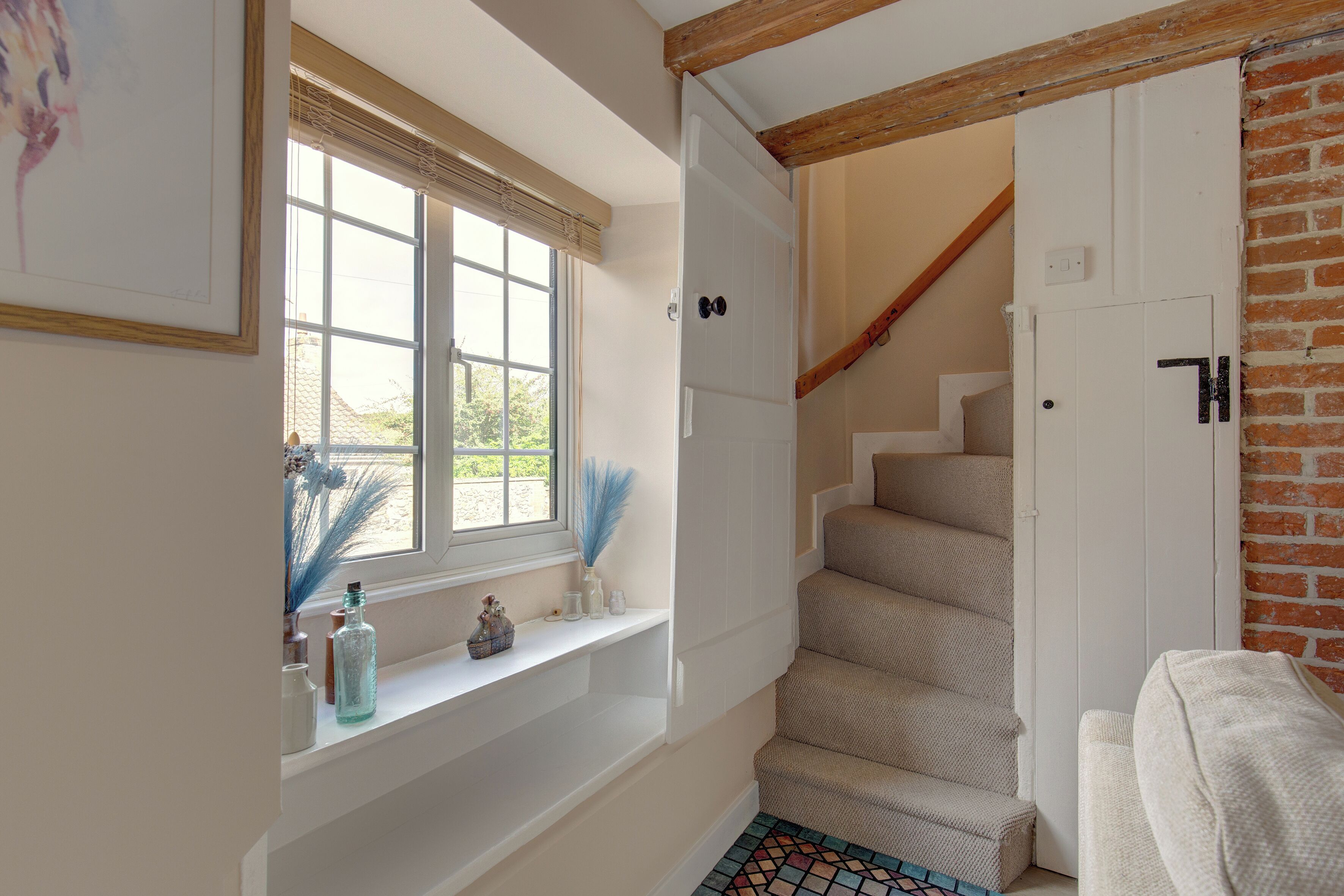 A staircase with a wooden beam ceiling, a white door, and a window with a view of greenery.
