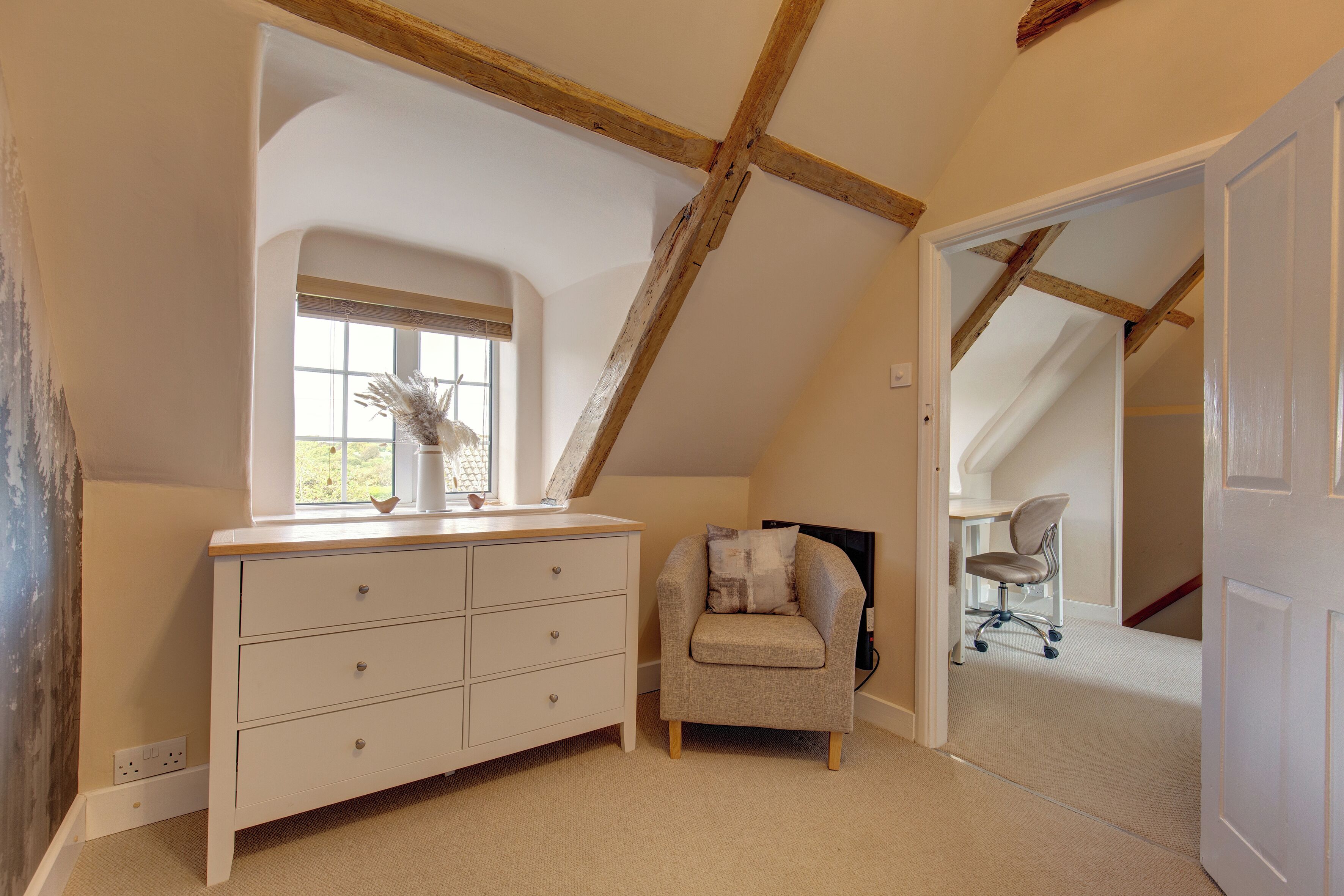 A room with a sloped ceiling, a white dresser, a grey armchair, and a window with a view.