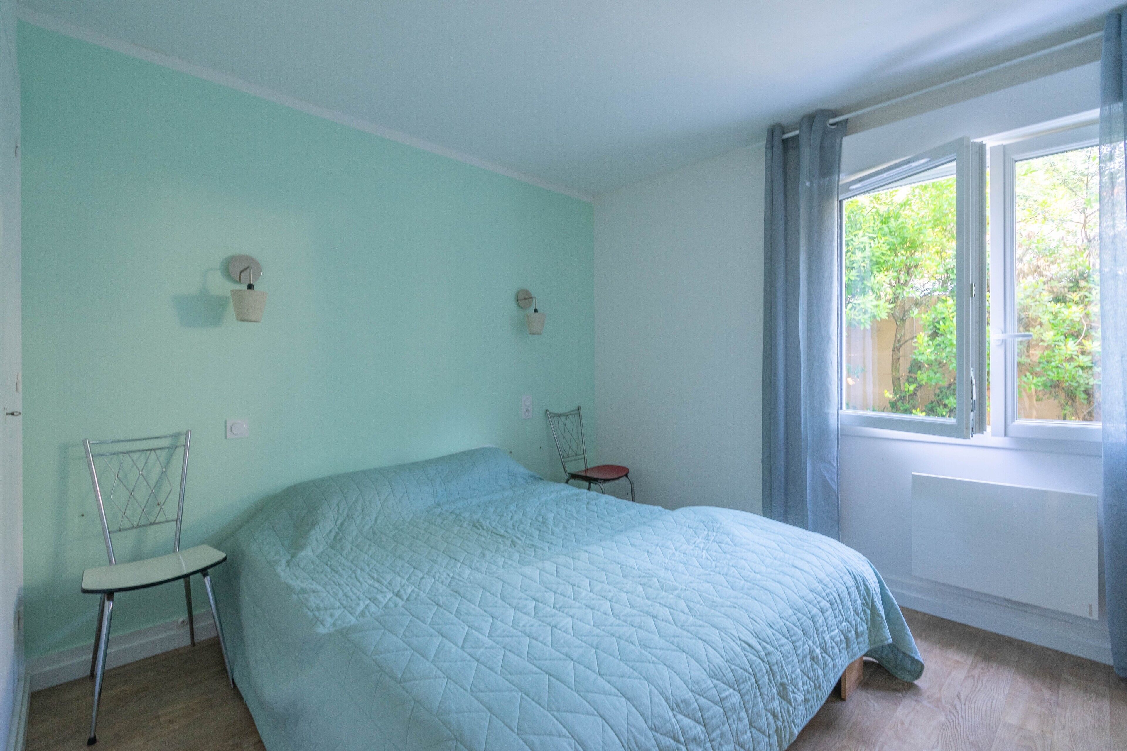 A bedroom with a bed, a chair, a window, and a wall-mounted light fixture.
